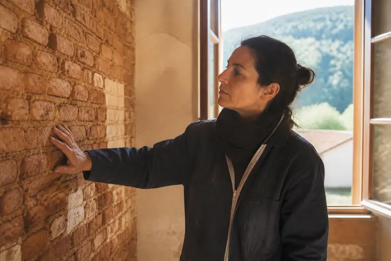 Une femme examine un mur en briques d'argile, soulignant le confort thermique et la régulation hygrométrique offerts par ce matériau traditionnel.